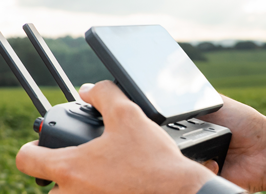 Operador segurando o controle de um drone agrícola New Holland, com o aplicativo de planejamento de voo na tela e o drone pulverizando a lavoura ao fundo, com o selo de Certificação CAAR New Holland