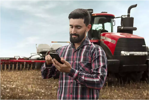 Agricultor sorrindo no campo em frente ao seu trator enquanto olha para seu celular e conferindo as melhorias de desempenho após aplicar a Consultoria de Alta Performance Case IH